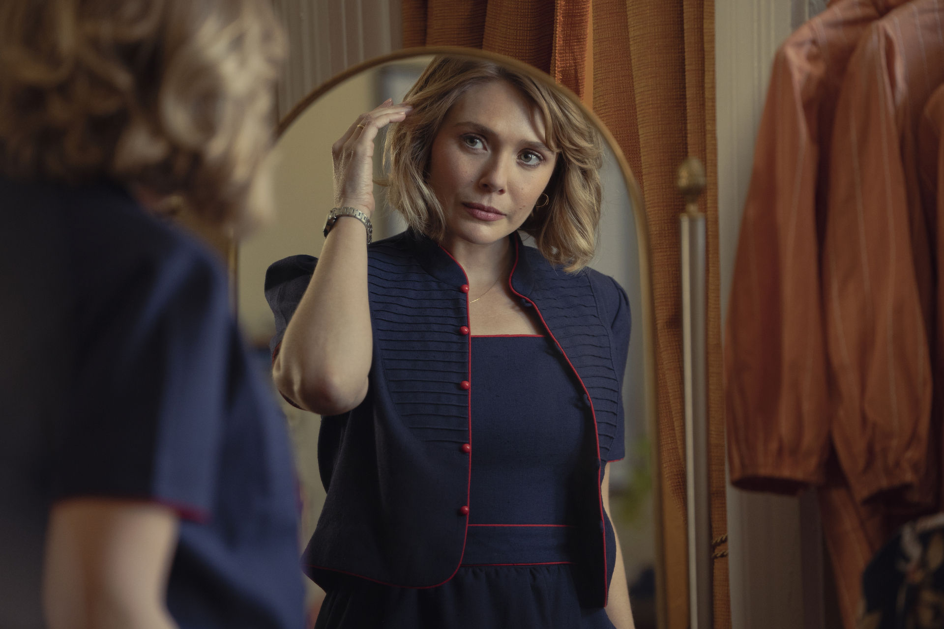 A woman with short, wavy blond hair wearing a navy blue dress and cover gazes at her reflection in the mirror; still of Elizabeth Olsen from "Love & Death"
