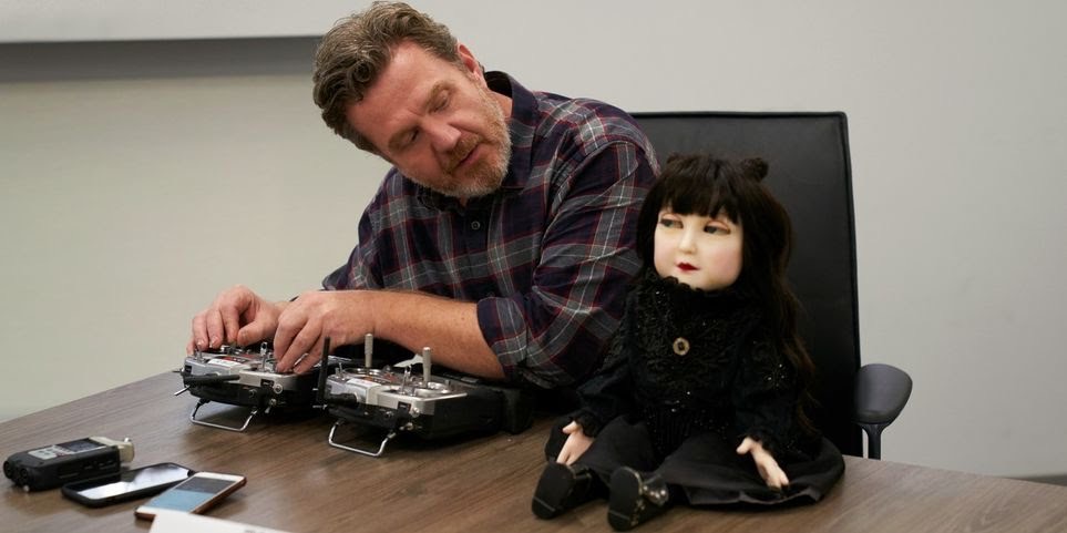 Animatronics designer Paul Jones sits at a table with Nadja doll sitting atop the table.  Jones is looking at the doll as he adjusts a remote control to move her head and eyes. 