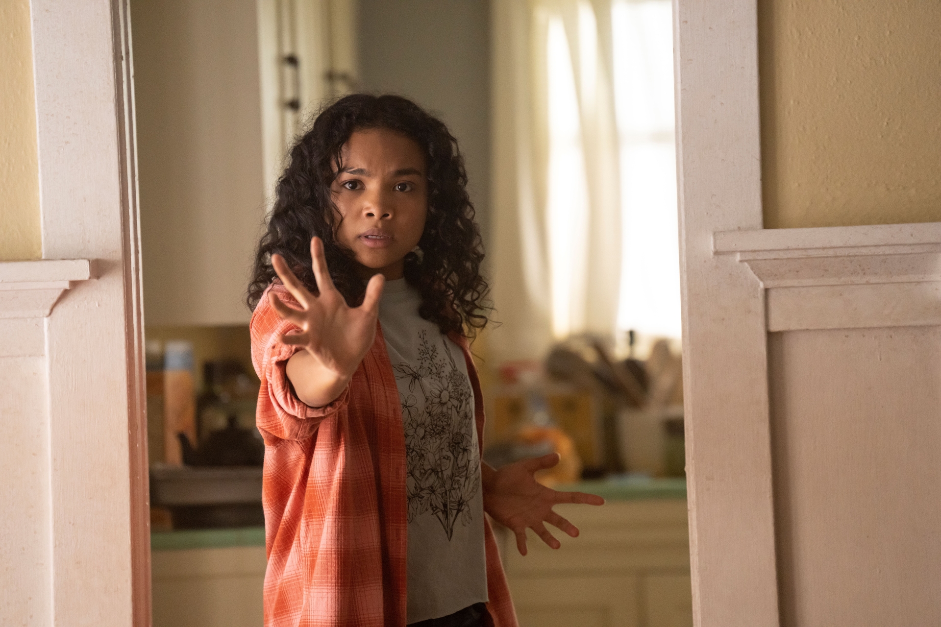 A teenage girl in a peach-and-pink plaid shirt and gray T-shirt stands just outside a kitchen, her hand outstretched in front of her to perform telekinetic tasks;  still from "Gray Matter"