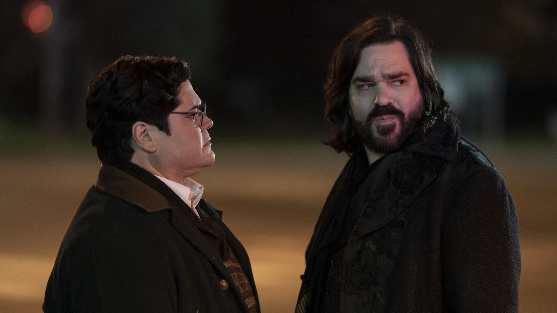 Medium shot of two men, one with glasses and the other with shoulder-length hair and a beard, speaking shadily at night; still from "What We Do in the Shadows"